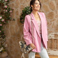 Woman in pink blazer and floral top with light blue denim jeans smiling while holding the Nicole Lee butterfly bag in gray around her shoulder to show varied carrying options set against a modern, rustic background with a bench and some flowers hanging behind her. 