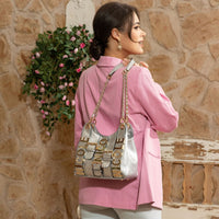 Woman in pink blazer holding the Nicole Lee Calista shoulder bag in metallic silver by the chain link and vegan leather dual straps around her shoulder set against a modern, rustic background with flowers hanging behind her.