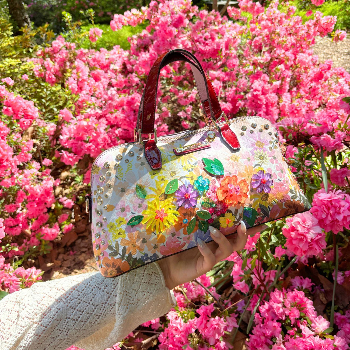 HOLDING OUR NICOLE LEE DOME HANDBAG IN DREAMS BLOSSOM DESIGN SET IN A GARDEN BACKGROUND.