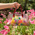 HOLDING OUR NICOLE LEE DOME HANDBAG IN DREAMS BLOSSOM DESIGN SET IN A GARDEN BACKGROUND.
