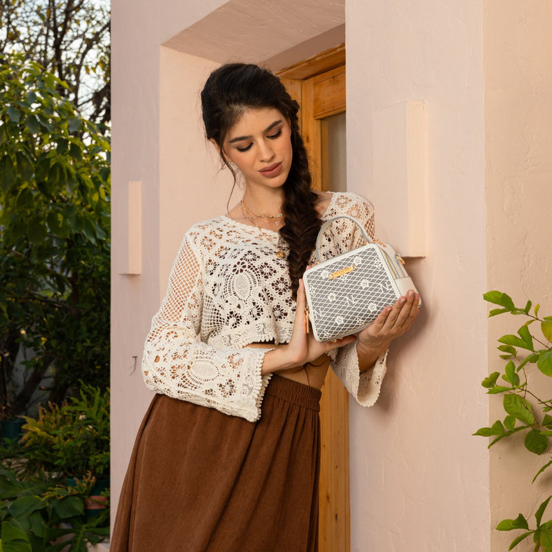 WOMEN IN A IVORY CROCKET CROP TOP AND BROWN SKIRT, LOOKING DOWN HOLDING OUR WHITE GRACIE MINI HANDBAG SET IN A MODERN BACKGROUND. 