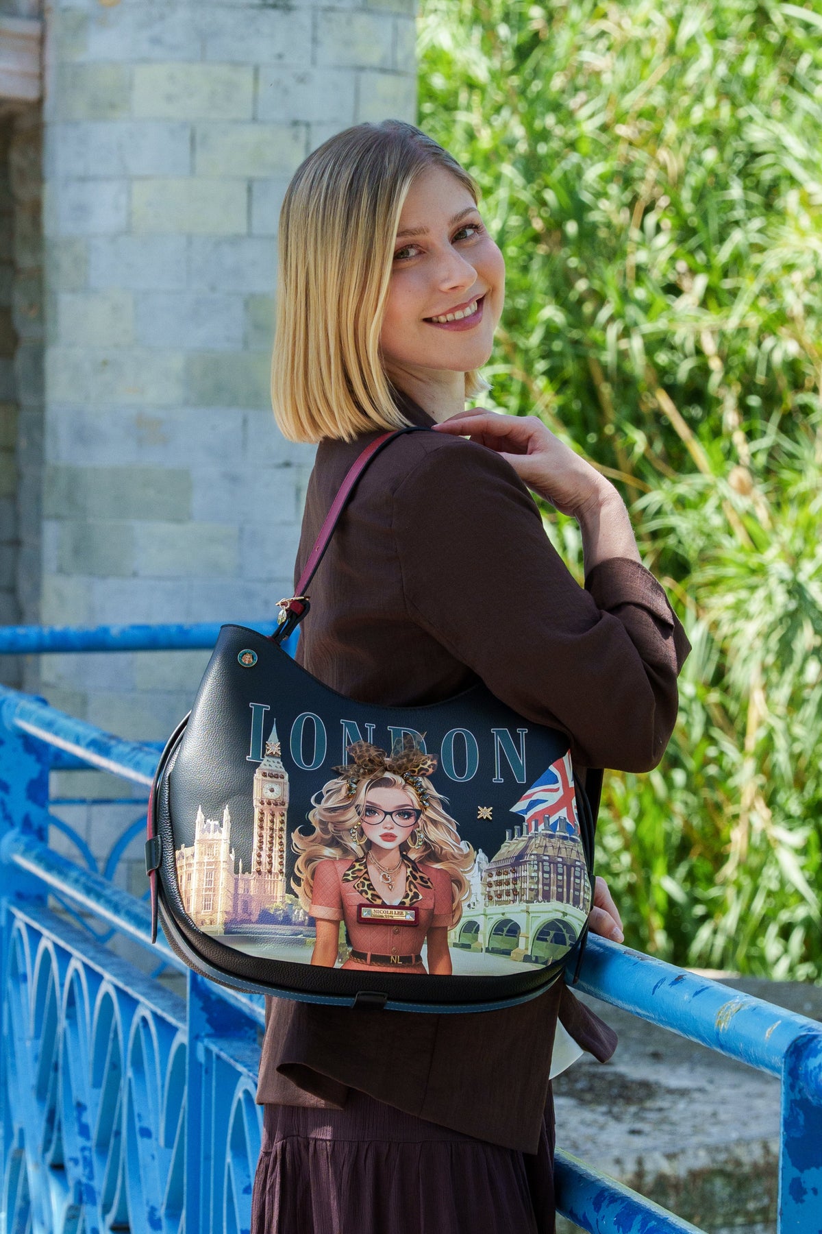 A woman with blonde hair stands on a bridge leaning on a blue railing, wearing a brown dress and carrying a black shoulder bag decorated with a London-themed illustration and the word 'LONDON'.