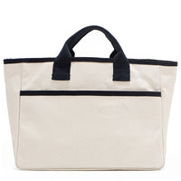 Back angle of the Emery handbag by Nodu Nadu in classic canvas-beige with navy blue trim. Made of polyester with dual top handles and a convenient black slip pocket to keep most-used items like your phone, keys, and planner close at hand. Dual top handles with a 3" drop allow you to carry this handbag by the top handles. 