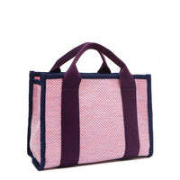Back angle view of the Nodu Nadu small canvas Marley tote Bag in pink. Accented with eggplant purple straps and a navy blue trim, this canvas features a subtle metallic shimmer that hits the light whenever you walk for a head-turning detail. 