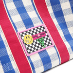 Close-up macro shot of the Nodu Nadu Blair Bag in blue with blue and white gingham print and hot pink shoulder strap accents. The front is adorned with an embroidered checker print stitch logo with baby pink trim and a smiley face and nodu nadu logo with USA flag.