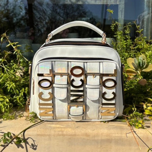Product shot of the Nicole Lee Calista top handle handbag in white with an iridescent white finishn on the buckle strap details with Nicole Lee Gold-tone hardware set against plants in a scenery outside.