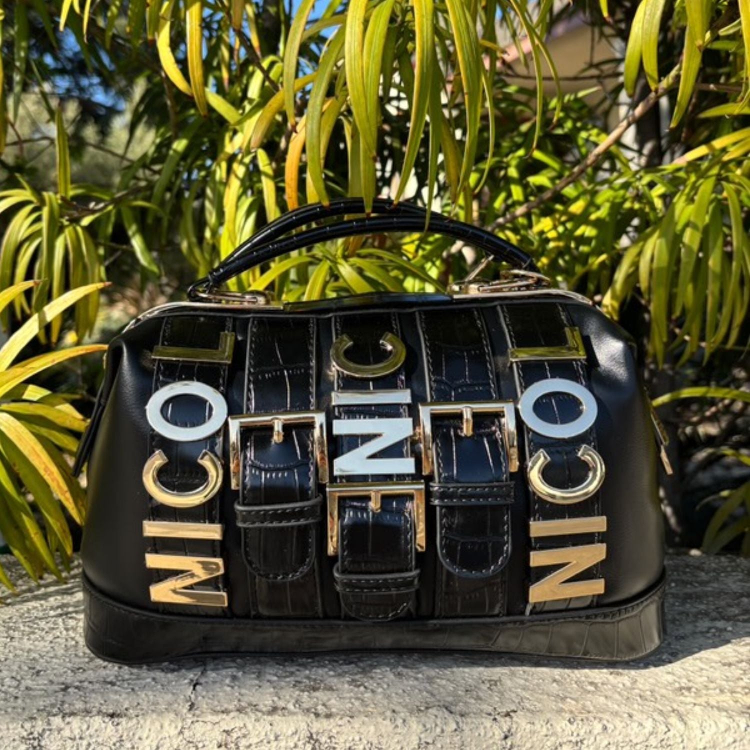 A lifestyle product shot of a black Nicole Lee Calista handbag with plants and foliage in the background. 