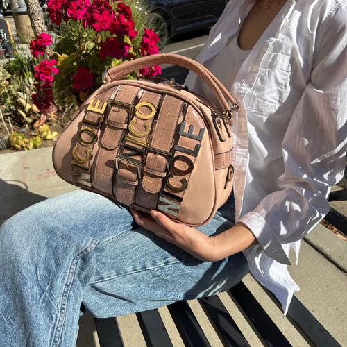 Cropped image of a woman holding a nude blush Nicole Lee Calista dome shaped handbag while sitting down on a bench outside on a sunny day with flowers in the background. 