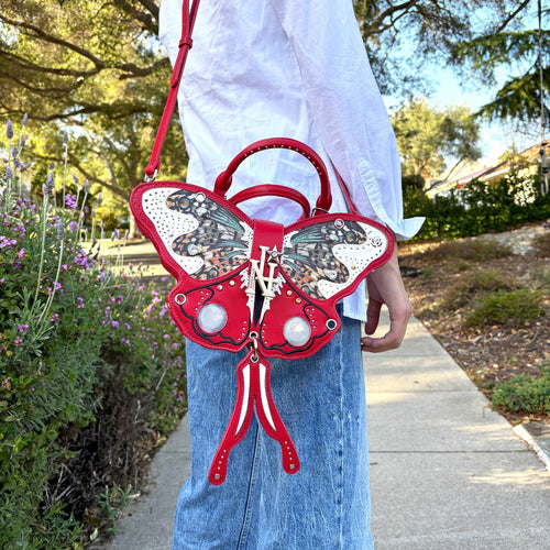 Woman wearing a white button up and blue denim pants carrying the Nicole Lee Butterfly Bag in red set against a sunny day in the neighborhood.